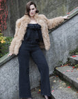 A woman posing on outdoor stone steps wearing a voluminous beige faux fur jacket over a black ruffled top and black wide-leg jeans.