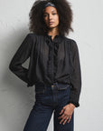 A woman modeling a black sheer long-sleeve blouse with ruffle trim and dark blue wide-leg jeans, standing against a light textured wall.