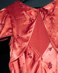 A close-up of a coral pink satin dress with a bow at the neckline, embellished with small red floral beadwork and rhinestones, hanging against a dark background.