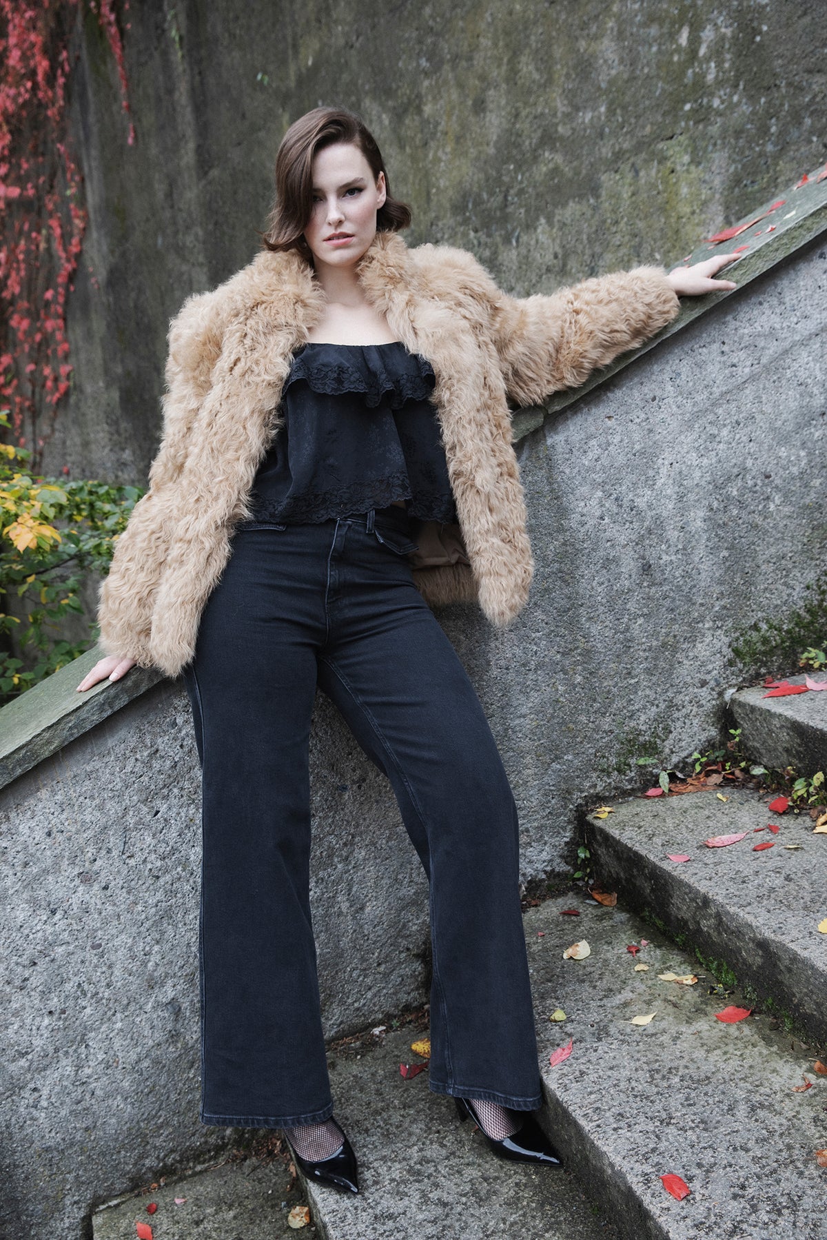 A woman posing on outdoor stone steps wearing a voluminous beige faux fur jacket over a black ruffled top and black wide-leg jeans.