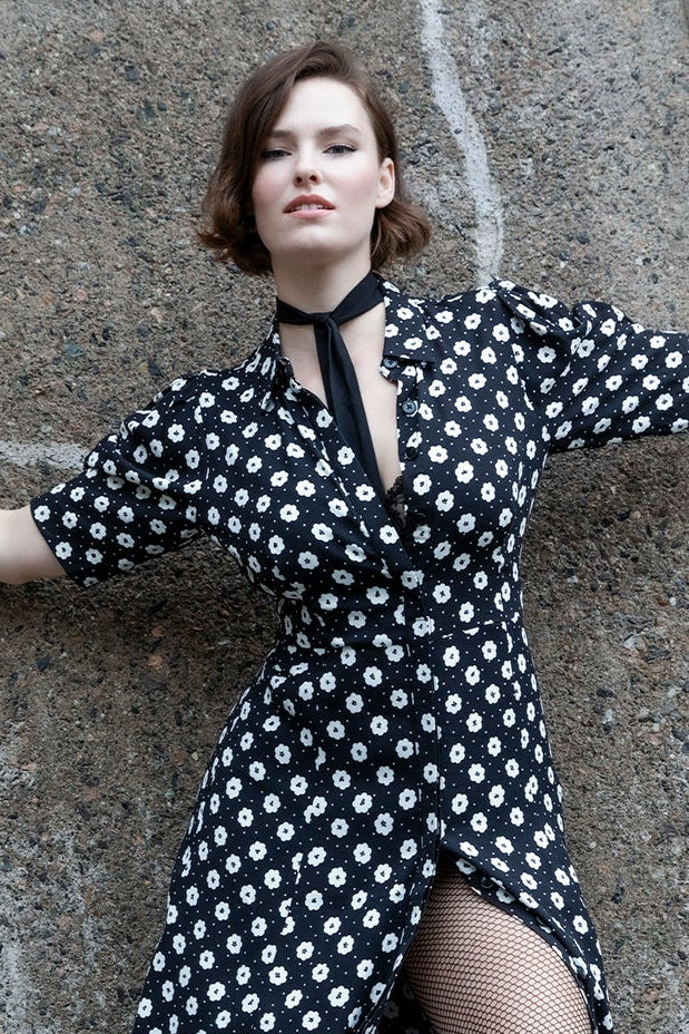 A woman posing in a black and white polka dot midi dress with a high-low hem and black tights, leaning against a textured concrete wall.