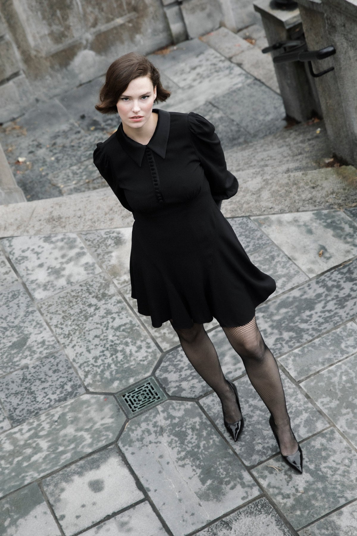 A woman wearing a black puff-sleeve mini dress, sheer black tights, and black pointed shoes stands on stone steps in an outdoor urban setting.