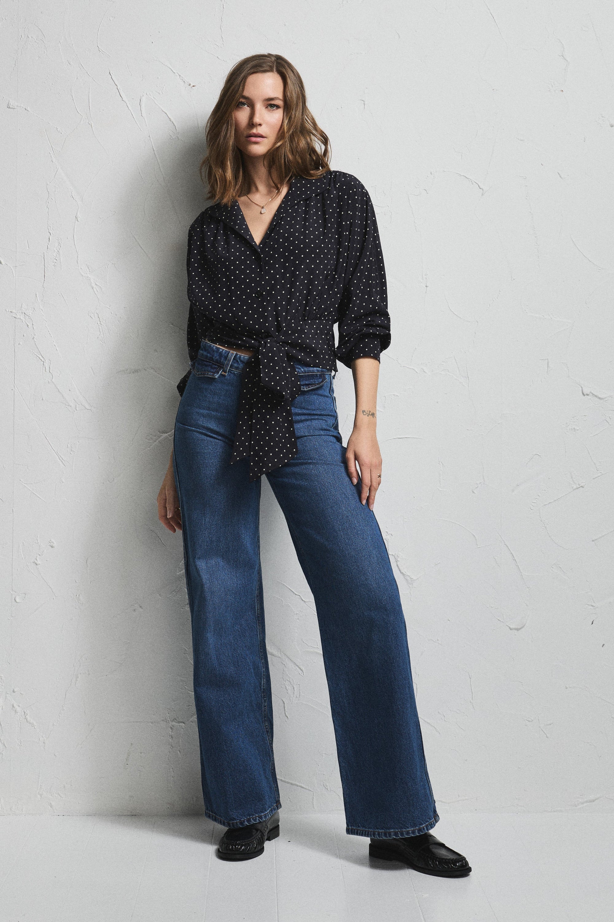 A woman stands against a textured white wall wearing a black and white patterned wrap-style blouse with three-quarter sleeves and blue wide-leg jeans.