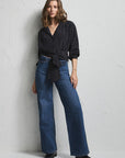A woman stands against a textured white wall wearing a black and white patterned wrap-style blouse with three-quarter sleeves and blue wide-leg jeans.