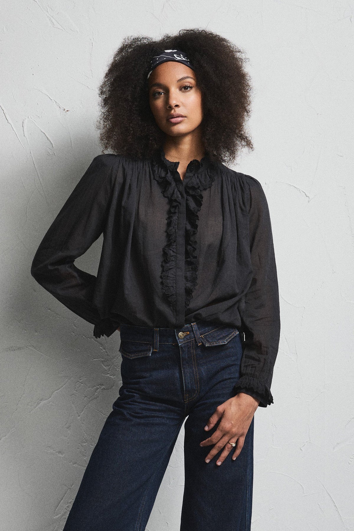 A woman modeling a black sheer long-sleeve blouse with ruffle trim and dark blue wide-leg jeans, standing against a light textured wall.