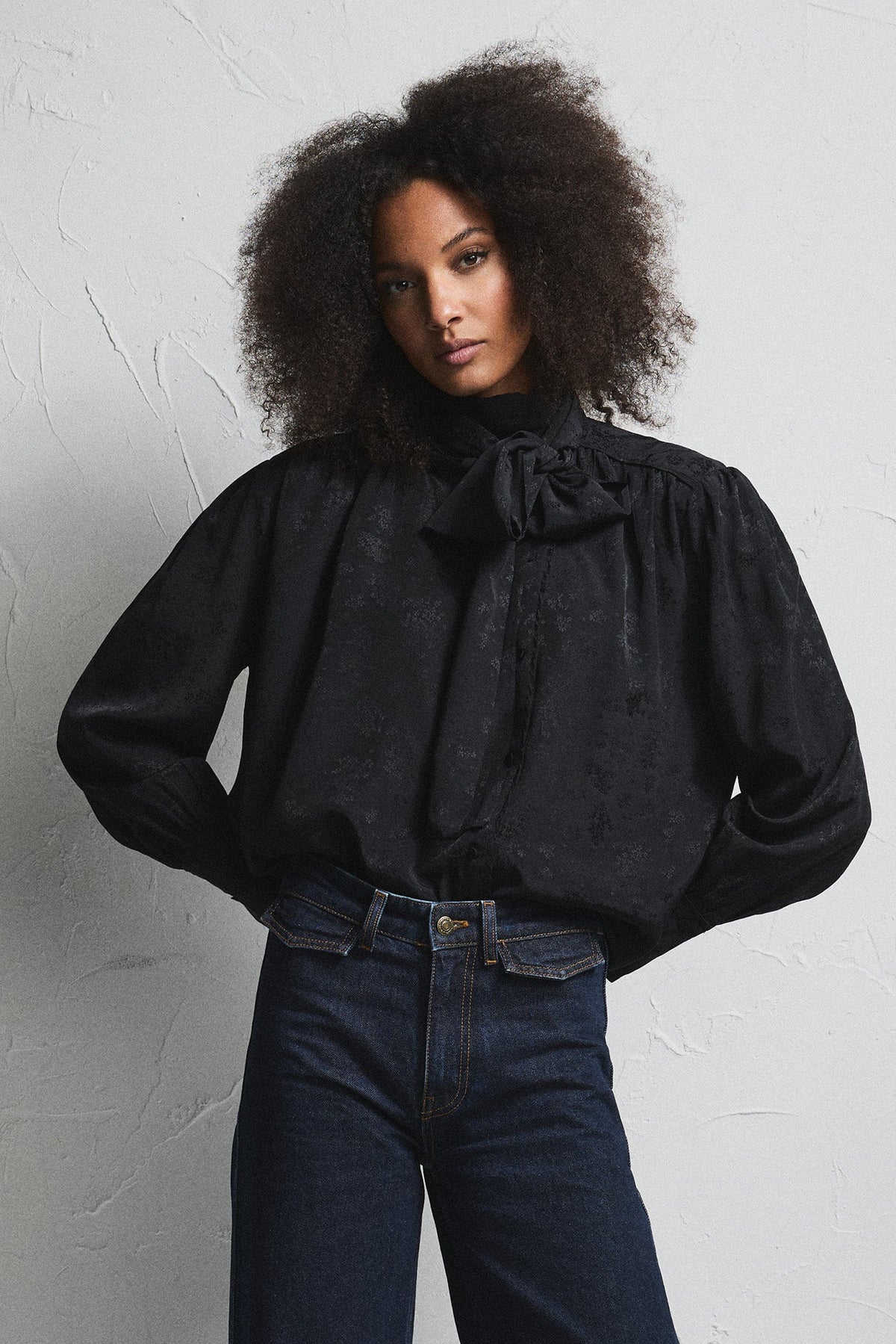 A model with curly dark hair stands against a textured white wall wearing a black long-sleeve blouse with a high ruffled collar and dark blue wide-leg jeans.