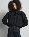 A model with curly dark hair stands against a textured white wall wearing a black long-sleeve blouse with a high ruffled collar and dark blue wide-leg jeans.