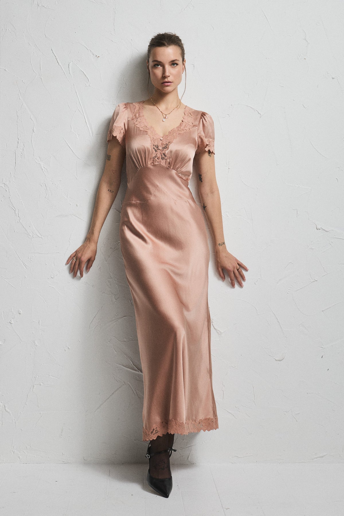 A woman stands against a textured white wall wearing a satin-like blush pink maxi dress with short flutter sleeves, lace trim at the neckline and hem, and black pointed shoes.