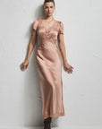 A woman stands against a textured white wall wearing a satin-like blush pink maxi dress with short flutter sleeves, lace trim at the neckline and hem, and black pointed shoes.