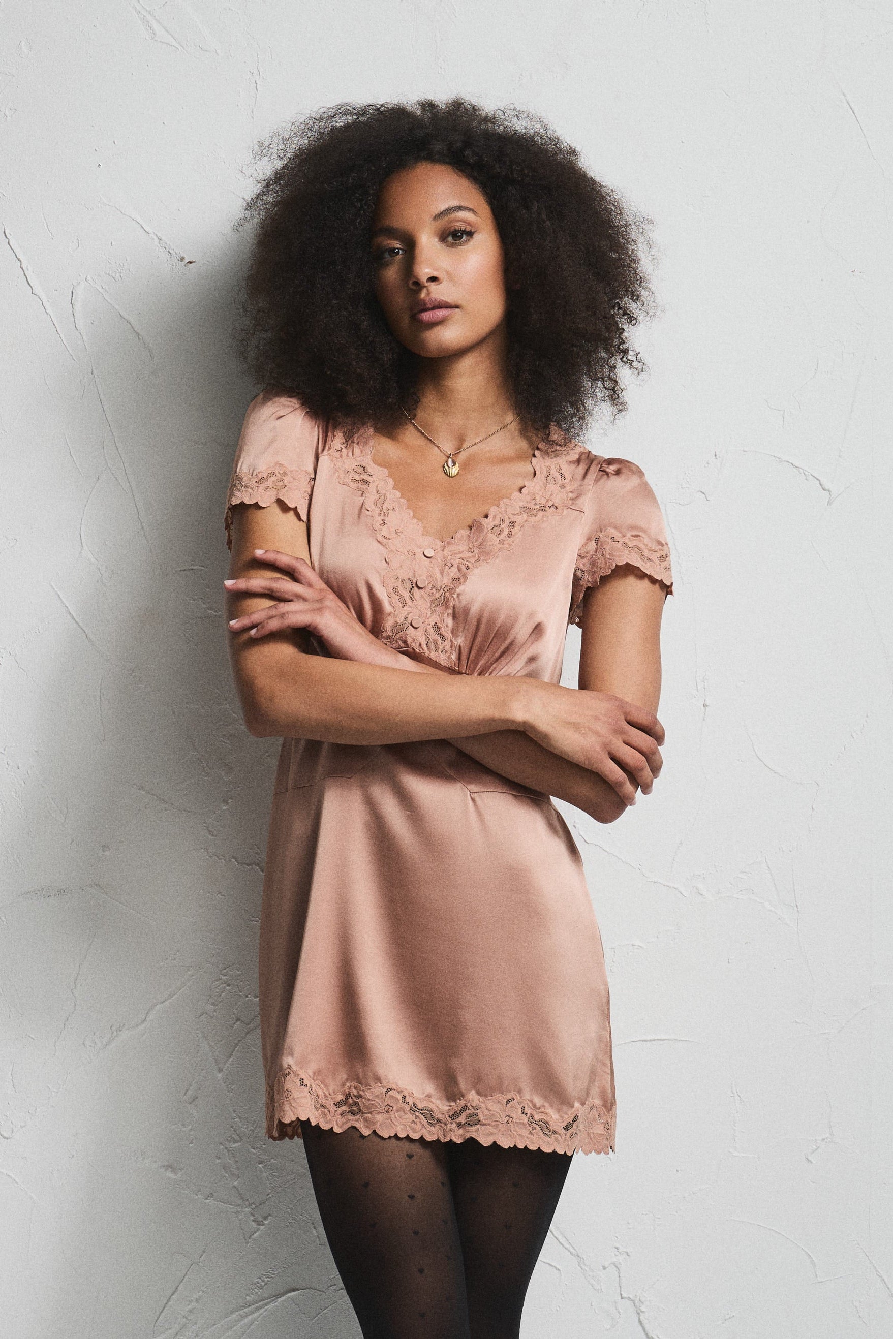 A woman with dark curly hair stands against a textured white wall wearing a short, shiny blush pink slip dress with lace trim, black tights, and black heels.