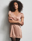A woman with dark curly hair stands against a textured white wall wearing a short, shiny blush pink slip dress with lace trim, black tights, and black heels.