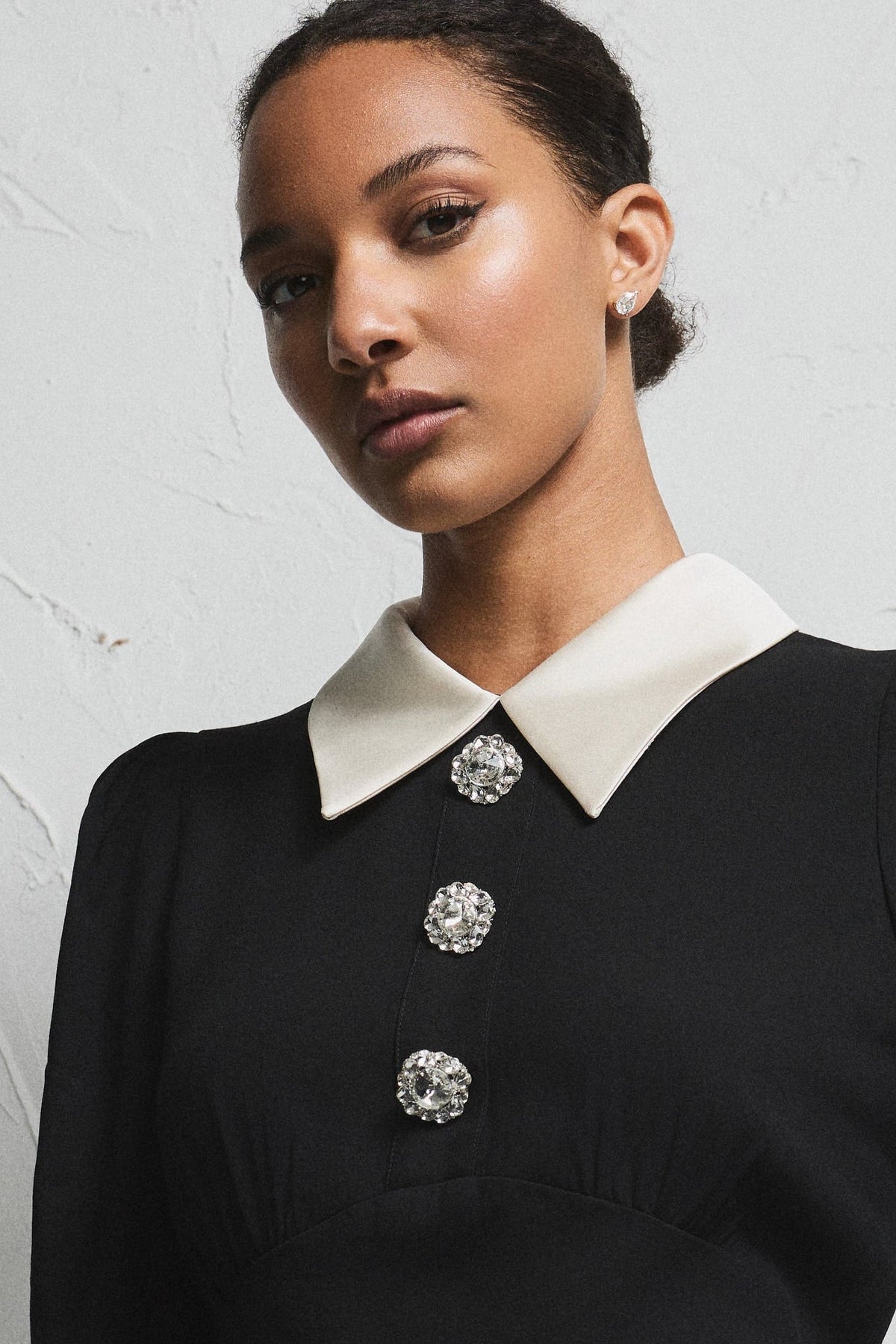 A model wearing a black long-sleeve peplum top with a contrasting white collar and cuffs, accented by three decorative silver buttons, paired with a matching black skirt, set against a textured light gray wall.