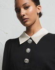 A model wearing a black long-sleeve peplum top with a contrasting white collar and cuffs, accented by three decorative silver buttons, paired with a matching black skirt, set against a textured light gray wall.