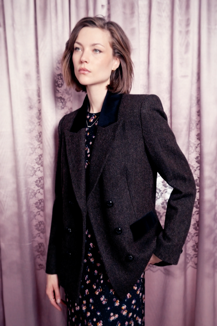 Woman wearing a dark coat over a patterned dress against a textured curtain background