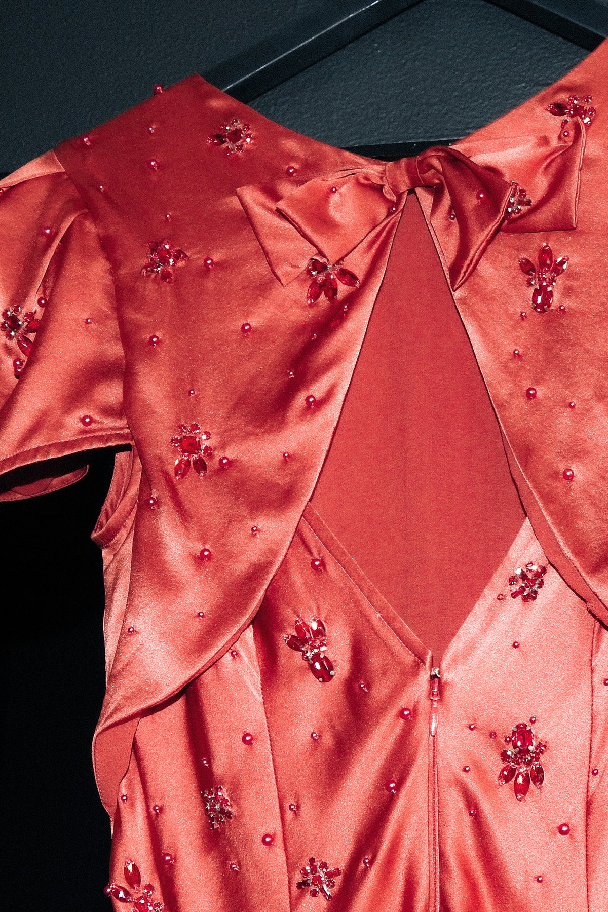 A close-up of a coral pink satin dress with a bow at the neckline, embellished with small red floral beadwork and rhinestones, hanging against a dark background.