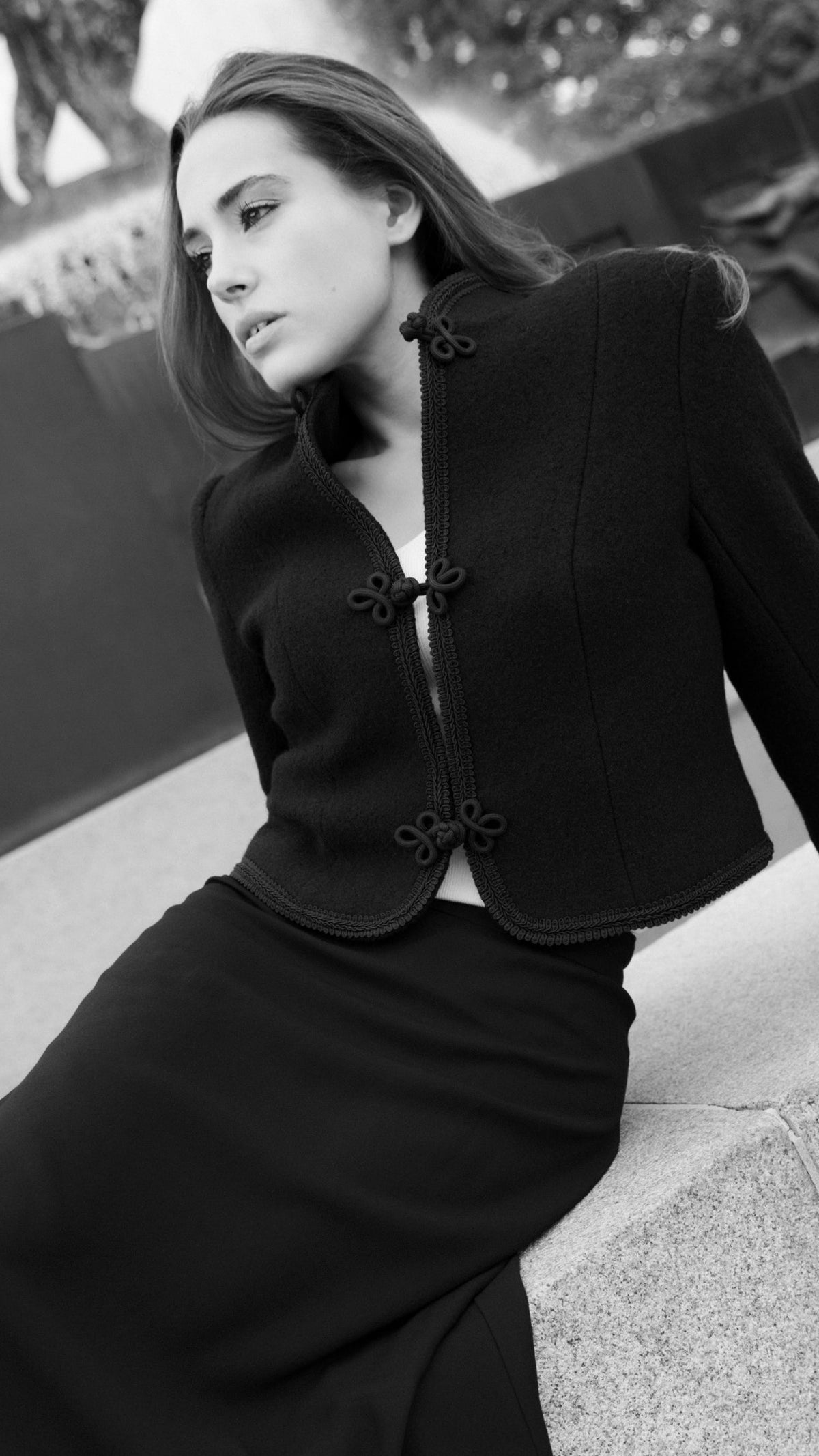 A woman wearing a black tailored jacket with decorative front buttons and a matching long skirt, posed on outdoor stone steps in a black-and-white photograph.