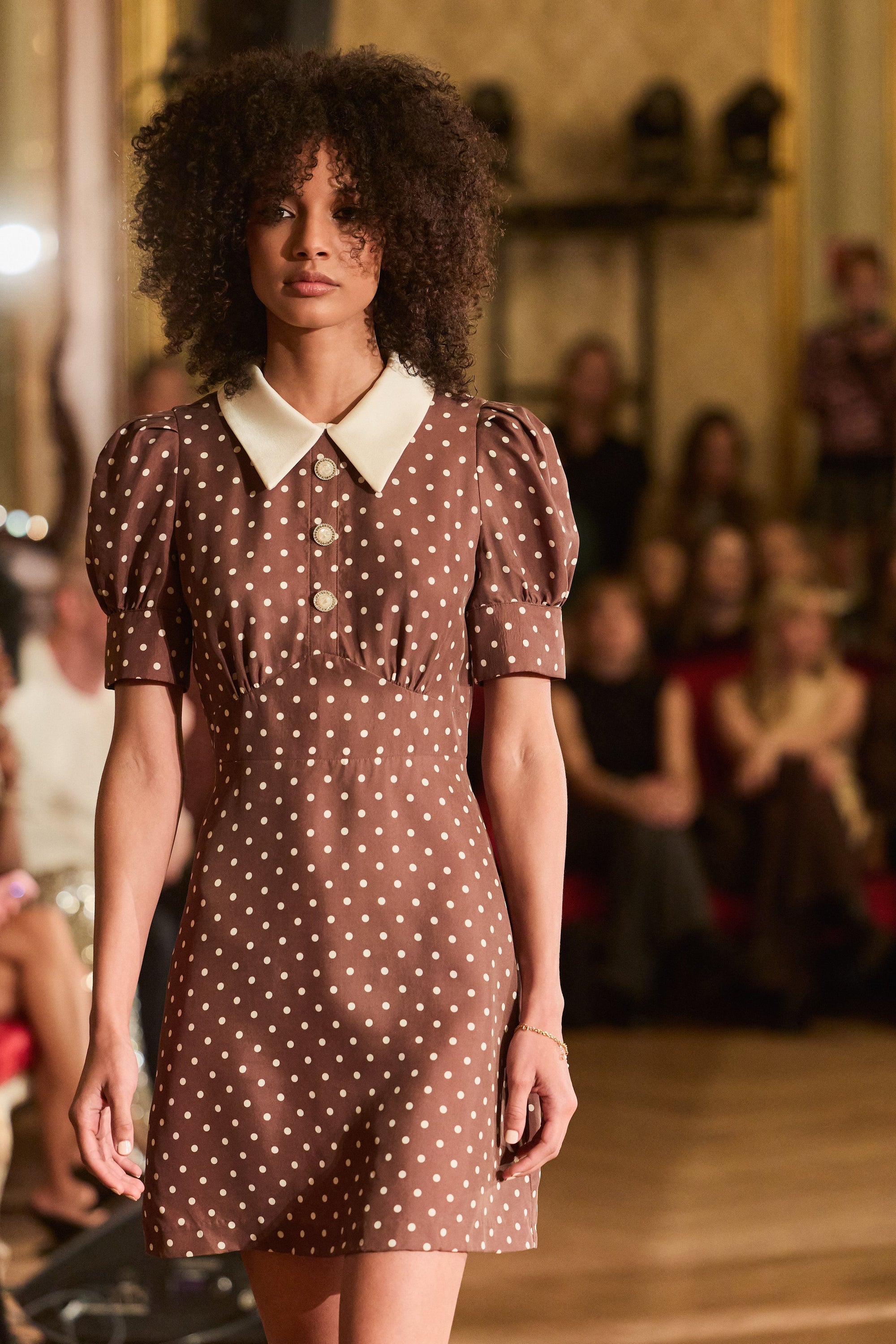 Model wearing a brown polka dot dress on a runway with an audience in the background.