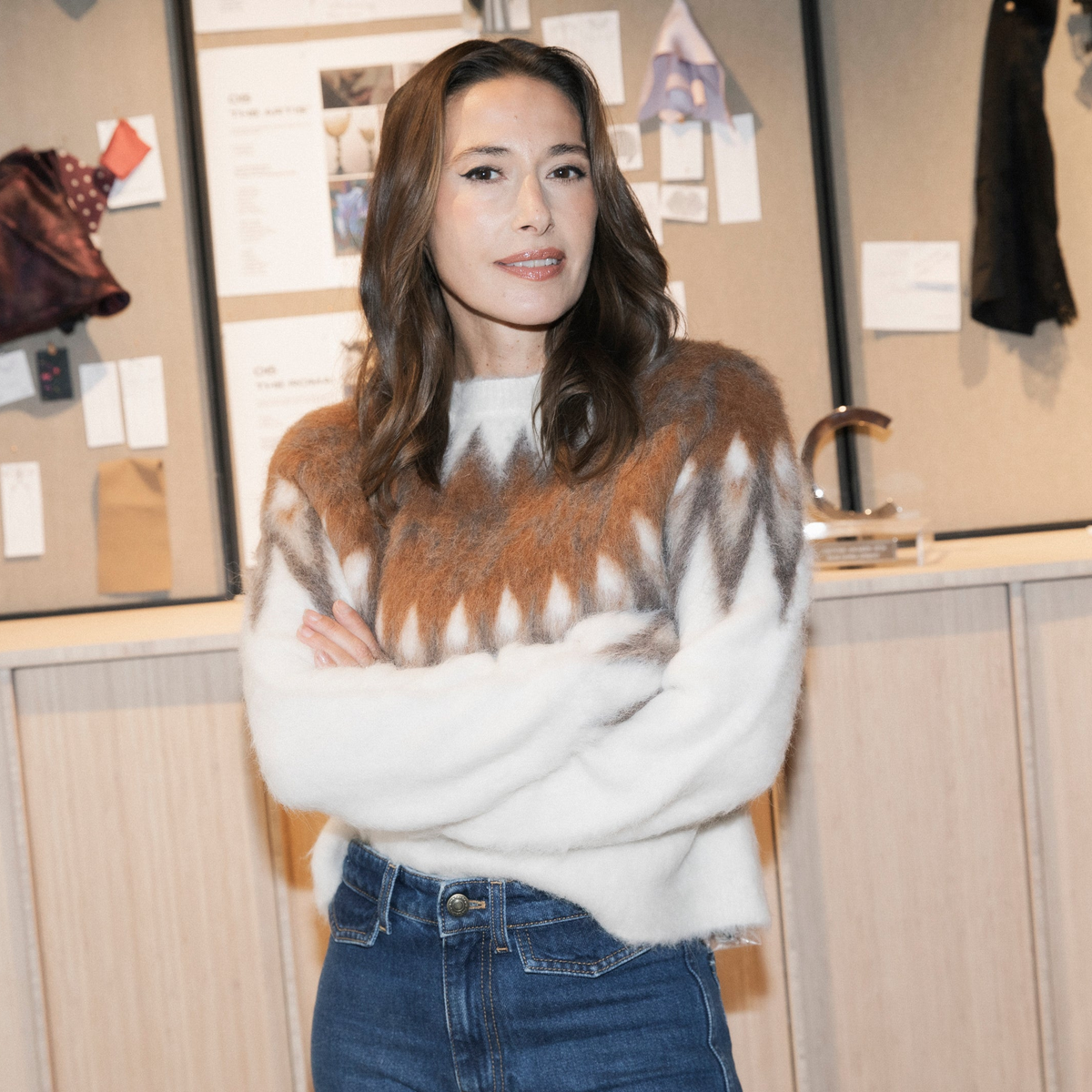 A woman with long brown hair stands with arms crossed, wearing a fuzzy knit sweater with a brown and white geometric pattern and blue jeans, set against an indoor studio or design office background.