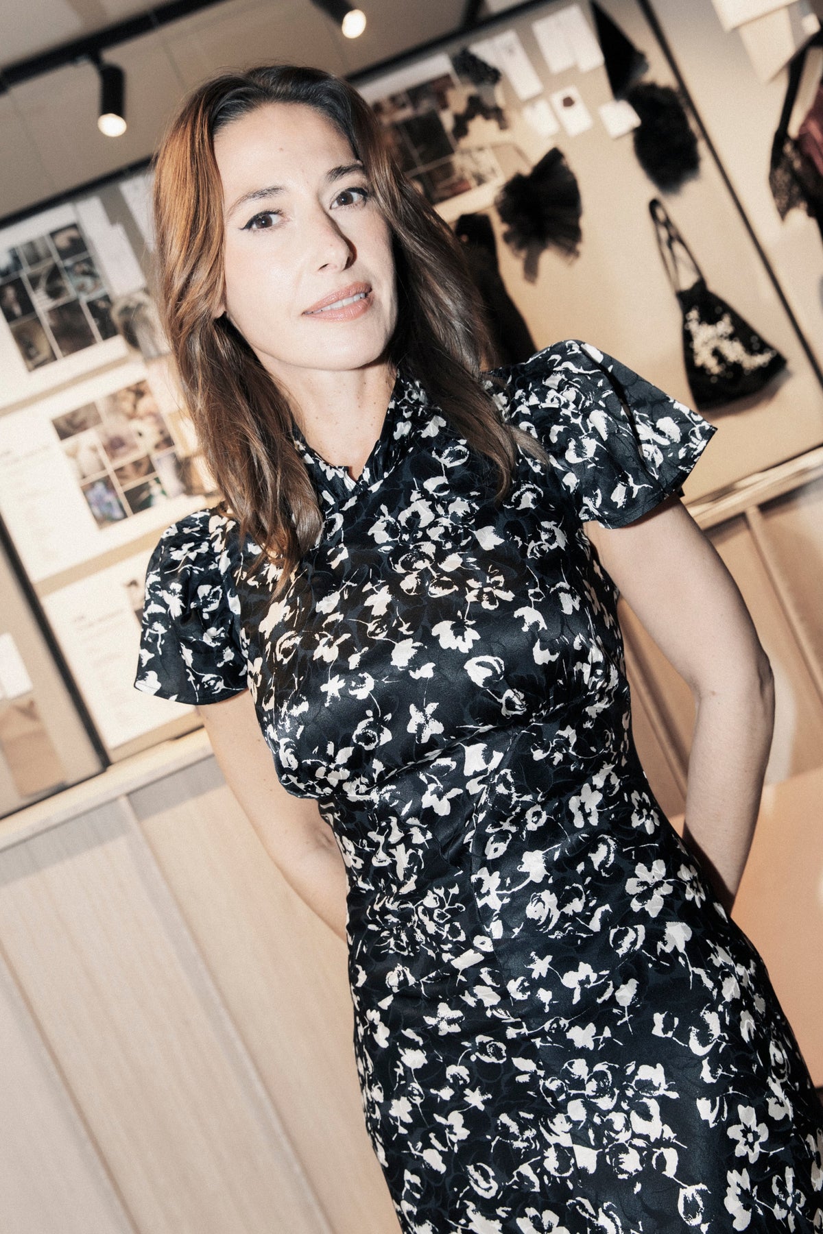 A woman wearing a black satin-like dress with a white floral print, standing in a boutique interior with framed fashion photos and bags in the background.