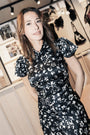 A woman wearing a black satin-like dress with a white floral print, standing in a boutique interior with framed fashion photos and bags in the background.
