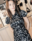 A woman wearing a black satin-like dress with a white floral print, standing in a boutique interior with framed fashion photos and bags in the background.