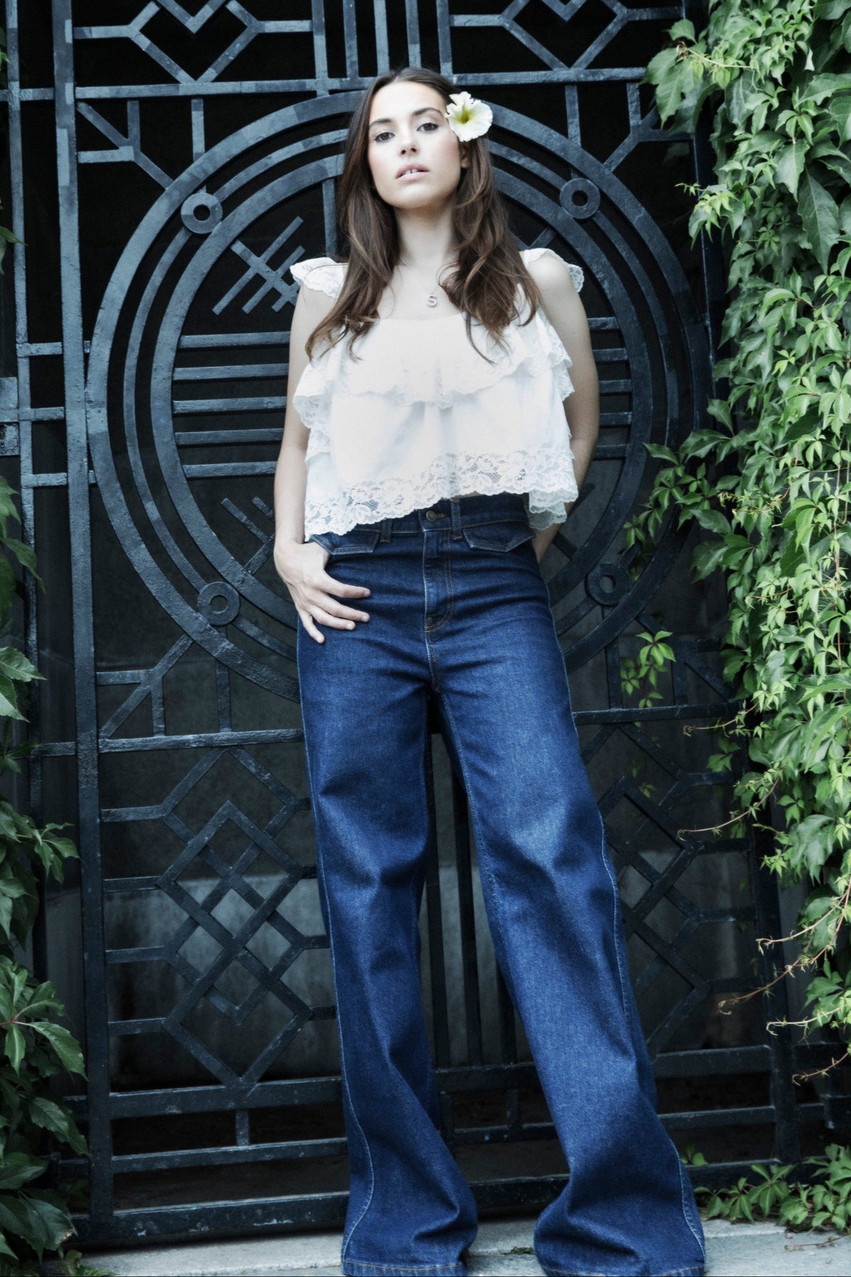 A woman stands in front of a black decorative metal gate surrounded by green ivy, wearing a white lace sleeveless top and high-waisted blue wide-leg jeans with a small white flower tucked behind her ear.