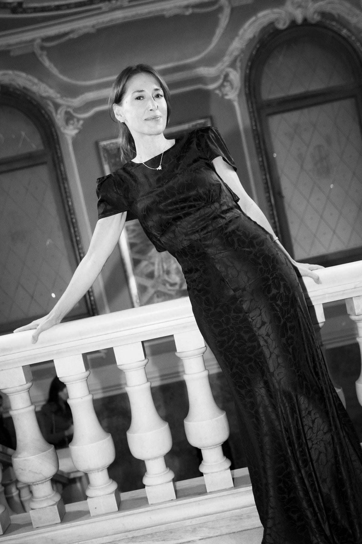 A woman wearing a black textured evening gown with short puff sleeves, leaning on a white stone balustrade in an ornate interior setting. The image is in black and white.