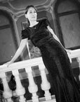 A woman wearing a black textured evening gown with short puff sleeves, leaning on a white stone balustrade in an ornate interior setting. The image is in black and white.
