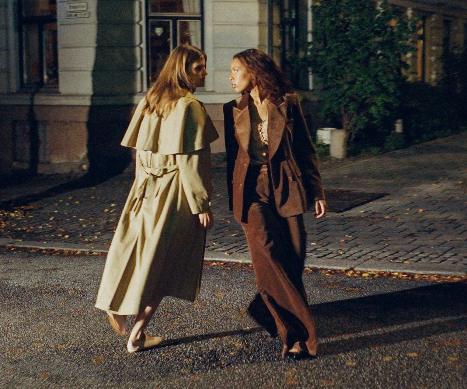 Two women walking and conversing on a city street at night. One wears a light beige trench coat, and the other wears a brown blazer and matching trousers.