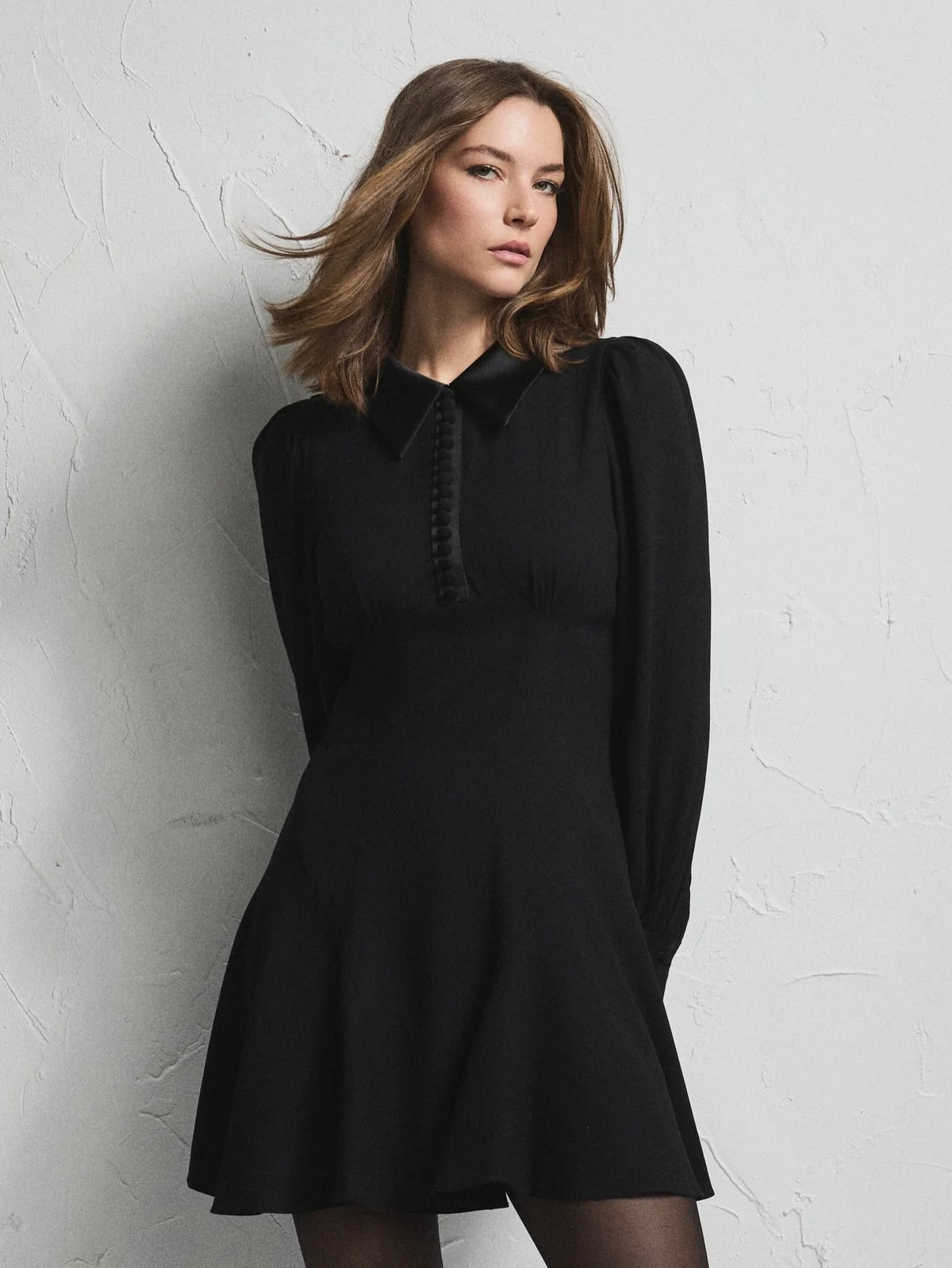 A woman wearing a black long-sleeve mini dress with a structured collar and decorative front detailing, posing against a textured light gray wall.